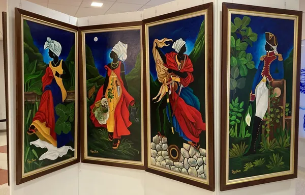 Four tall painted panels displayed as a folding screen: three women in head wraps and flowing red, yellow and blue dresses in a lush green landscape, and a figure in a Haitian revolutionary officer’s uniform and bicorne