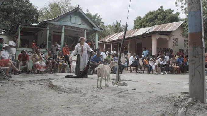 A singer in a long white costume performs on a dirt road beside a goat, watched by villagers seated in front of houses with tin roofs
