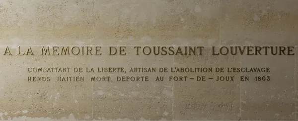 Gilded capitals on a stone wall: “A la mémoire de Toussaint Louverture, combattant de la liberté, artisan de l’abolition de l’esclavage, héros haïtien mort déporté au Fort-de-Joux en 1803”